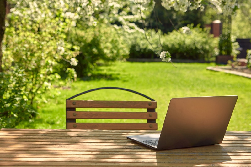 A laptop with stands on a wooden table in the courtyard of a beautiful home garden, in Sunny weather