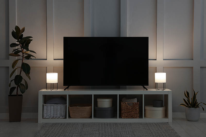 Modern TV on cabinet, lamps and beautiful houseplants near white wall in room